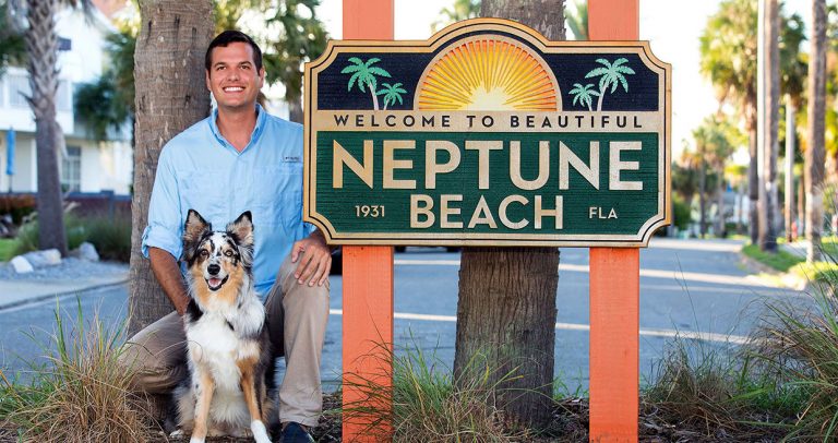 Josh Messinger kneels next to the Neptune Beach City sign with a brown, white and black dog with a happy face and large ears