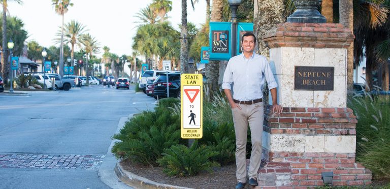 Josh Messinger leaning against a column with a plague saying Neptune beach on a corner of a shopping and roadway area